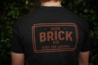 Person wearing a black t-shirt with 'Suck Brick Kid Stay the Course' text on the back, standing against a green leafy background.