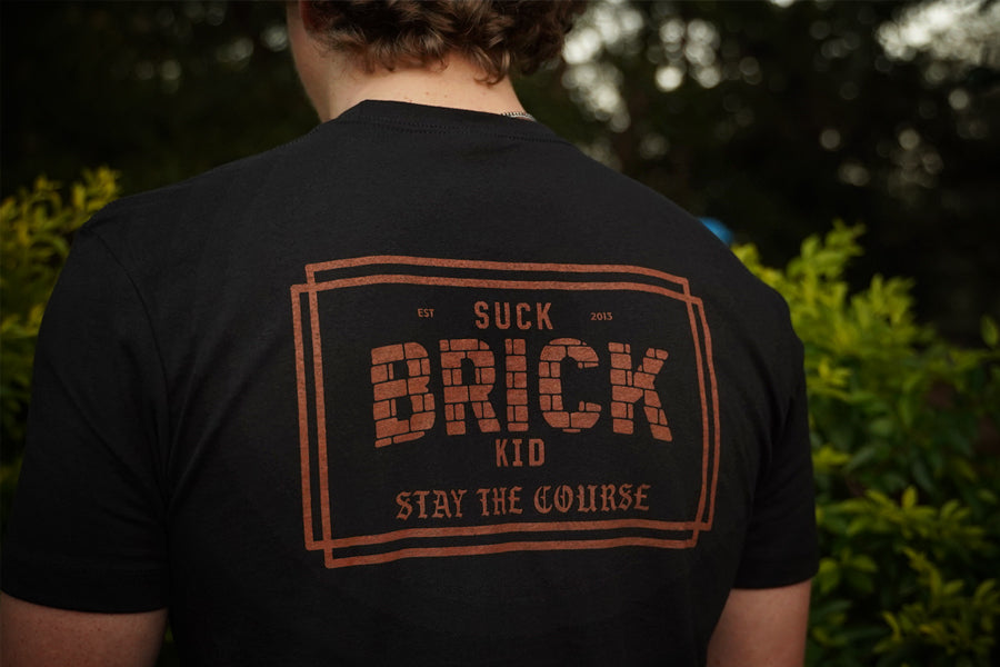 Person wearing a black t-shirt with 'SUCK BRICK KID' text on the back, outdoors.