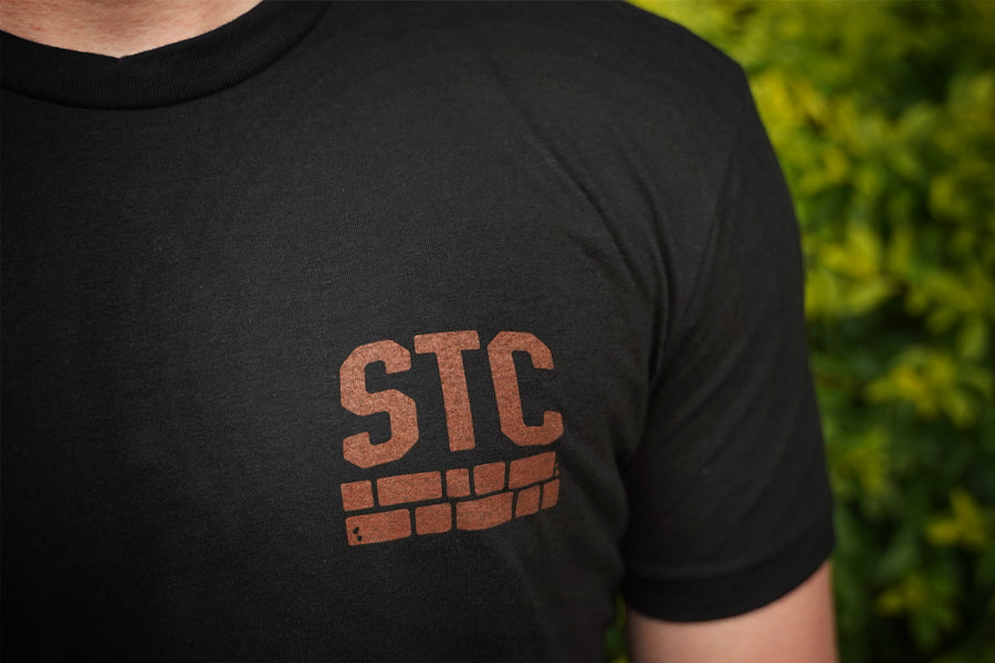 Black t-shirt with 'STC' logo worn by a person, against a blurred green background