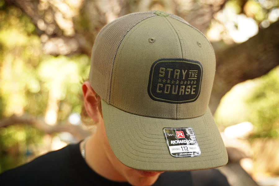 Olive cap with 'Stay the Course' embroidery worn by a person outdoors