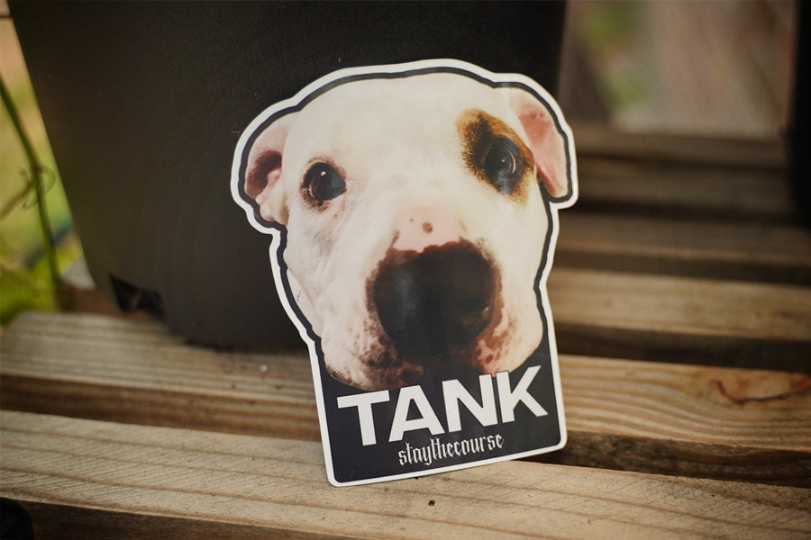 Sticker of a dog's face with 'TANK' and 'staythecourse' text on a wooden surface.
