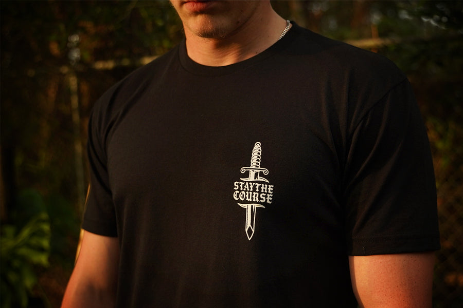 A person wearing a black t-shirt with what print of dagger and Stay the Course on the chest, in an outdoor setting