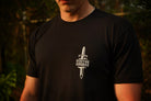 A person wearing a black t-shirt with what print of dagger and Stay the Course on the chest, in an outdoor setting