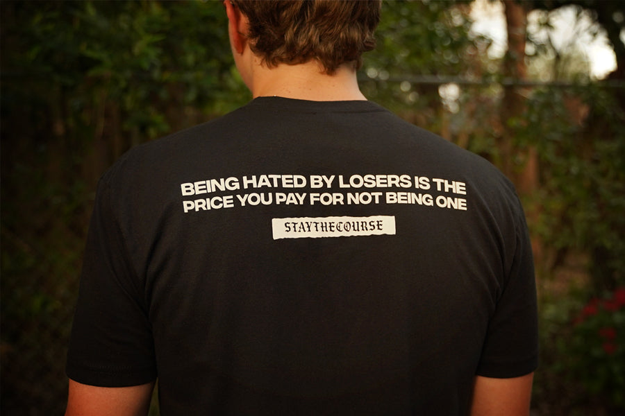Person wearing a black t-shirt with white print that says "Being hated by losers is the price you pay for not being one, Stay the Course", in an outdoor setting