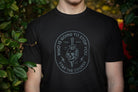 Person wearing a black t-shirt with a graphic design and text, standing against a natural background.