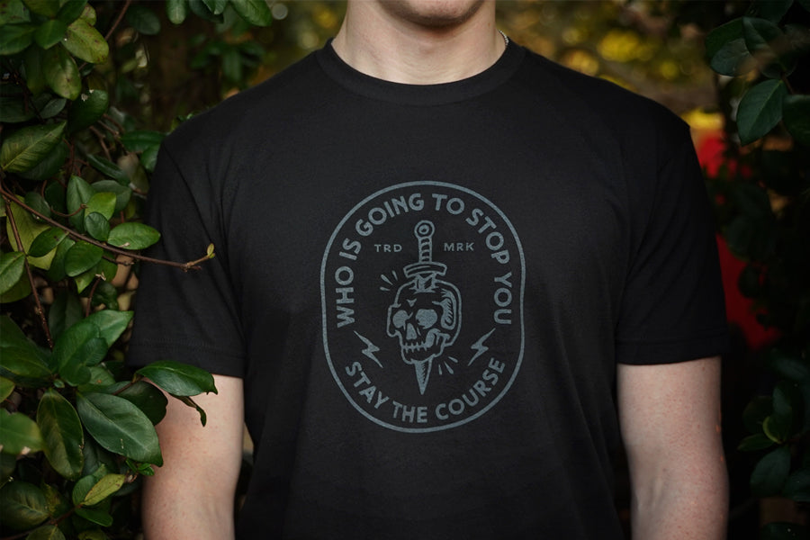 Person wearing a black t-shirt with a graphic design and text, standing against a natural background.