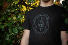 Person wearing a black t-shirt with a graphic design in front of green foliage