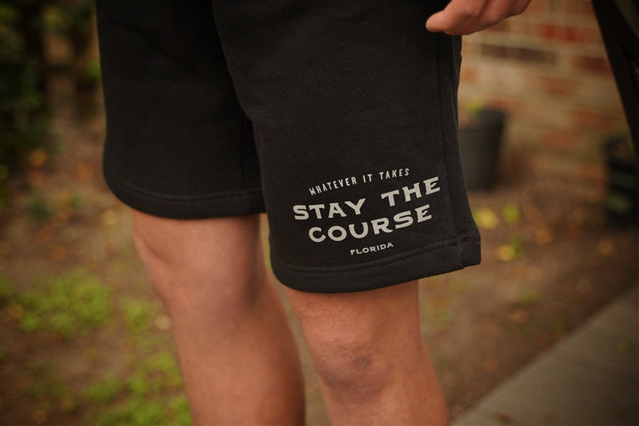 Black shorts with text 'Stay the Course Florida' held by a person outdoors.