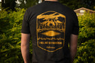 Person wearing a black t-shirt with a gold graphic and text in an outdoor setting